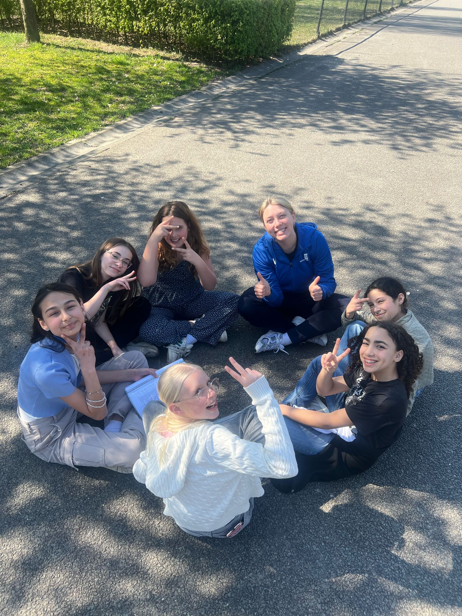 De zon schijnt, in een kring zitten een aantal verschillende meiden samen. Ze kijken in de camera en lachen. Bij hen in de cirkel zit een sportprofessional.